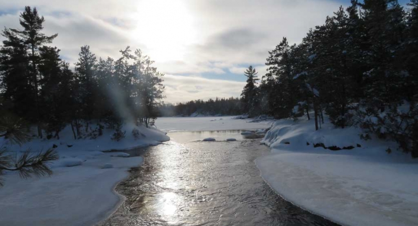 A creek passes through a snowy landscape and is framed on either side by trees. 